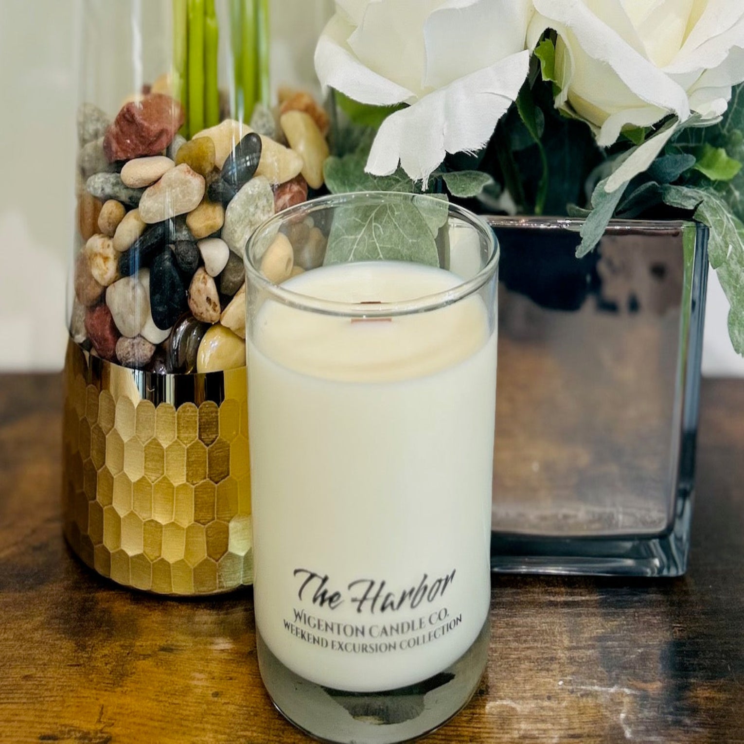 Candle with 'The Harbor' label in front of decorative vases with stones and flowers on a wooden surface