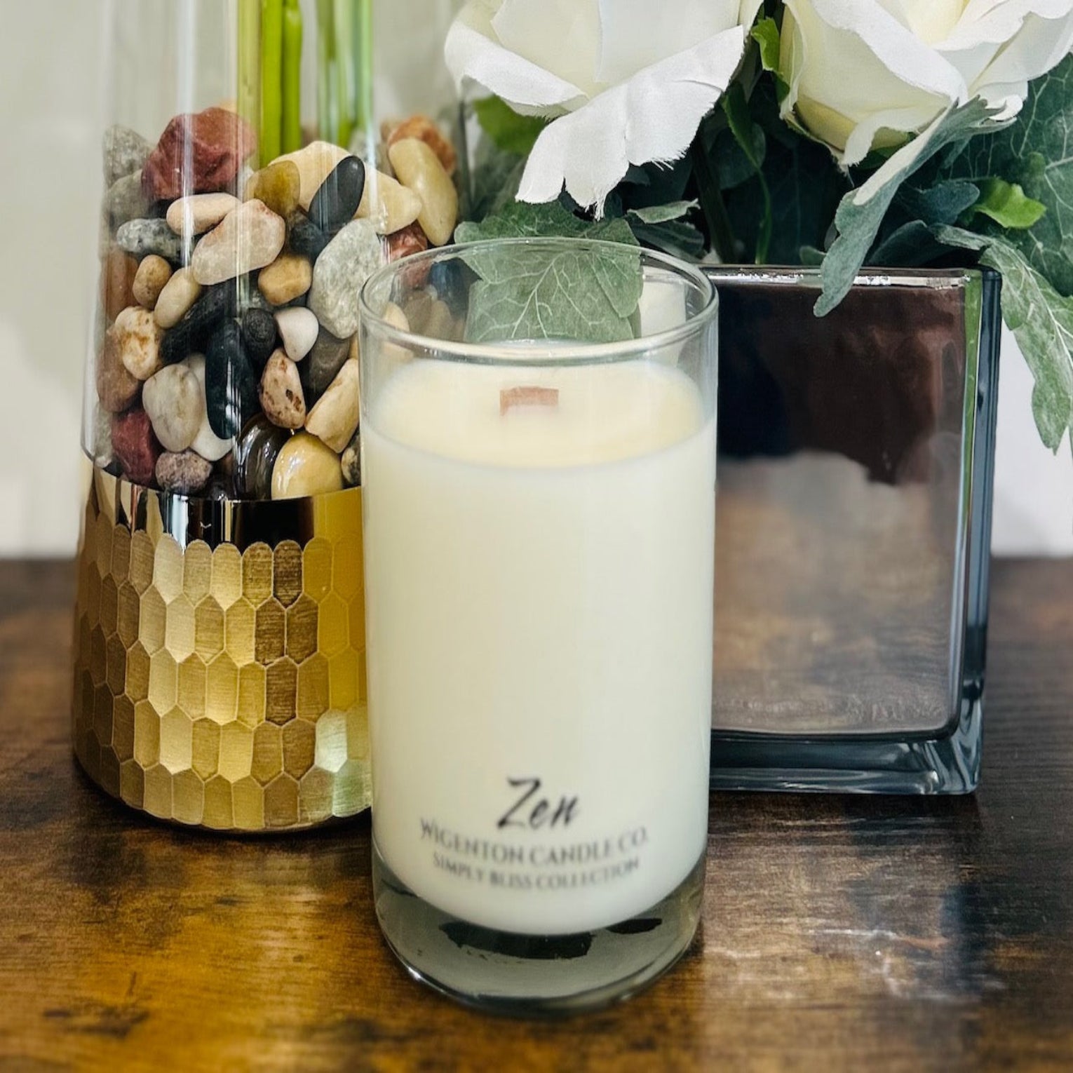 Candle labeled 'Zen' by Wigenton Candle Co. with decorative stones and flowers in the background.