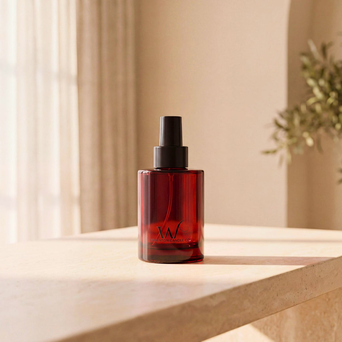 Wigenton Candle Co. 3.4 oz fragrance mist in an amber glass bottle with a black spray pump, on a sunlit ivory stone table with soft curtains and greenery in the background.