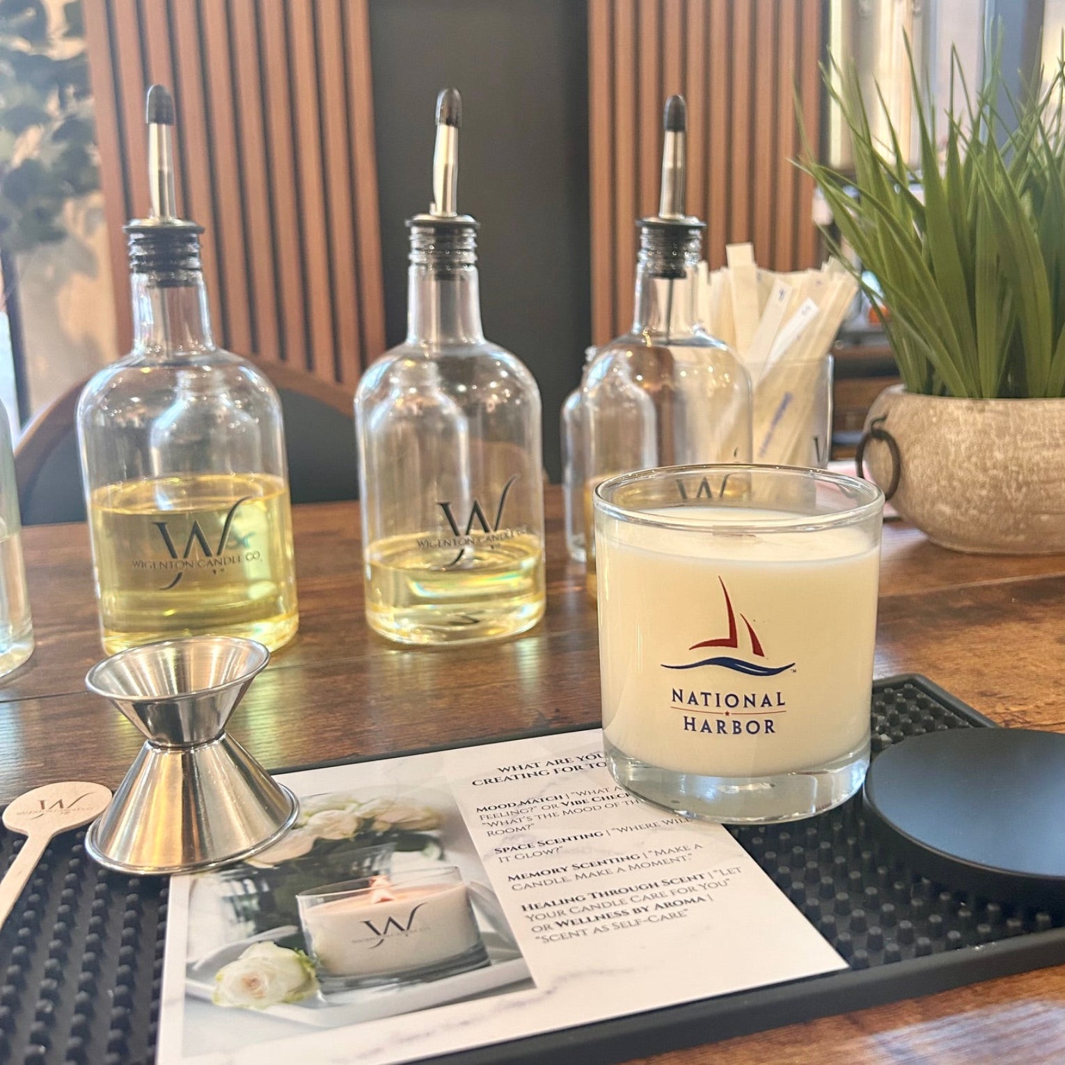 Candle labeled 'National Harbor' on a table with bottles and a plant in the background. Fragrance card underneath. 