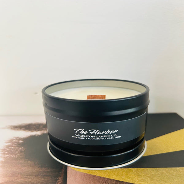 Candle in a black tin labeled 'The Harbor' by Wigenton Candle Co. on a wooden surface with a white background.