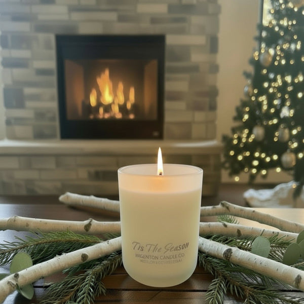 ’Tis the Season candle centered on a wooden table in front of a fireplace and Christmas tree, framed closely with warm lighting for a cozy and balanced presentation