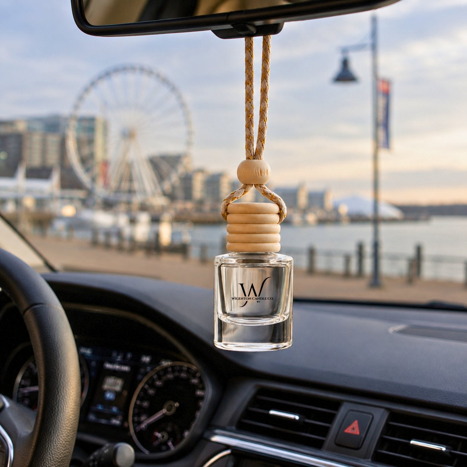 Hanging car diffuser by Wigenton Candle Co. featuring a wooden cap and glass fragrance vessel with National Harbor waterfront visible through the windshield.