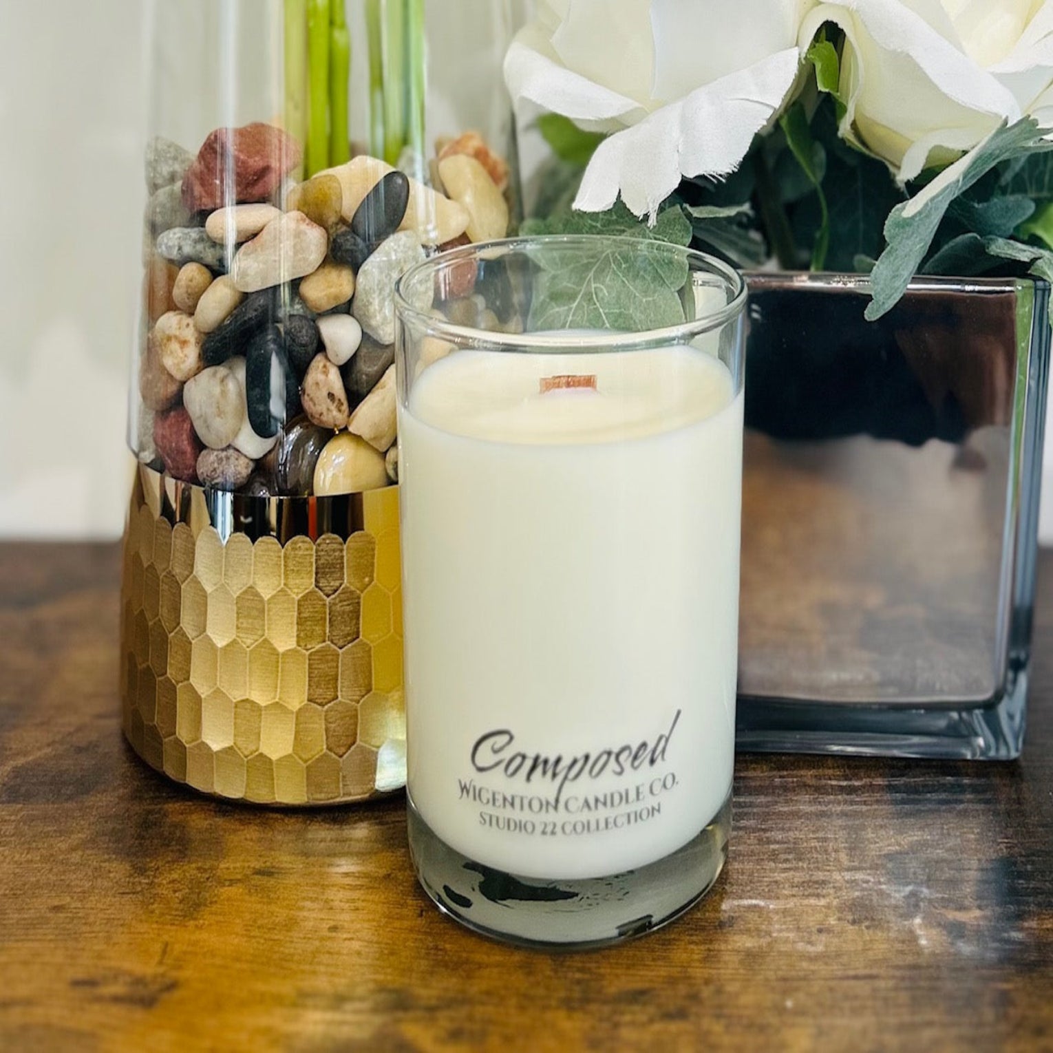 Glass candle with 'Composed' branding in front of decorative vases on a wooden surface