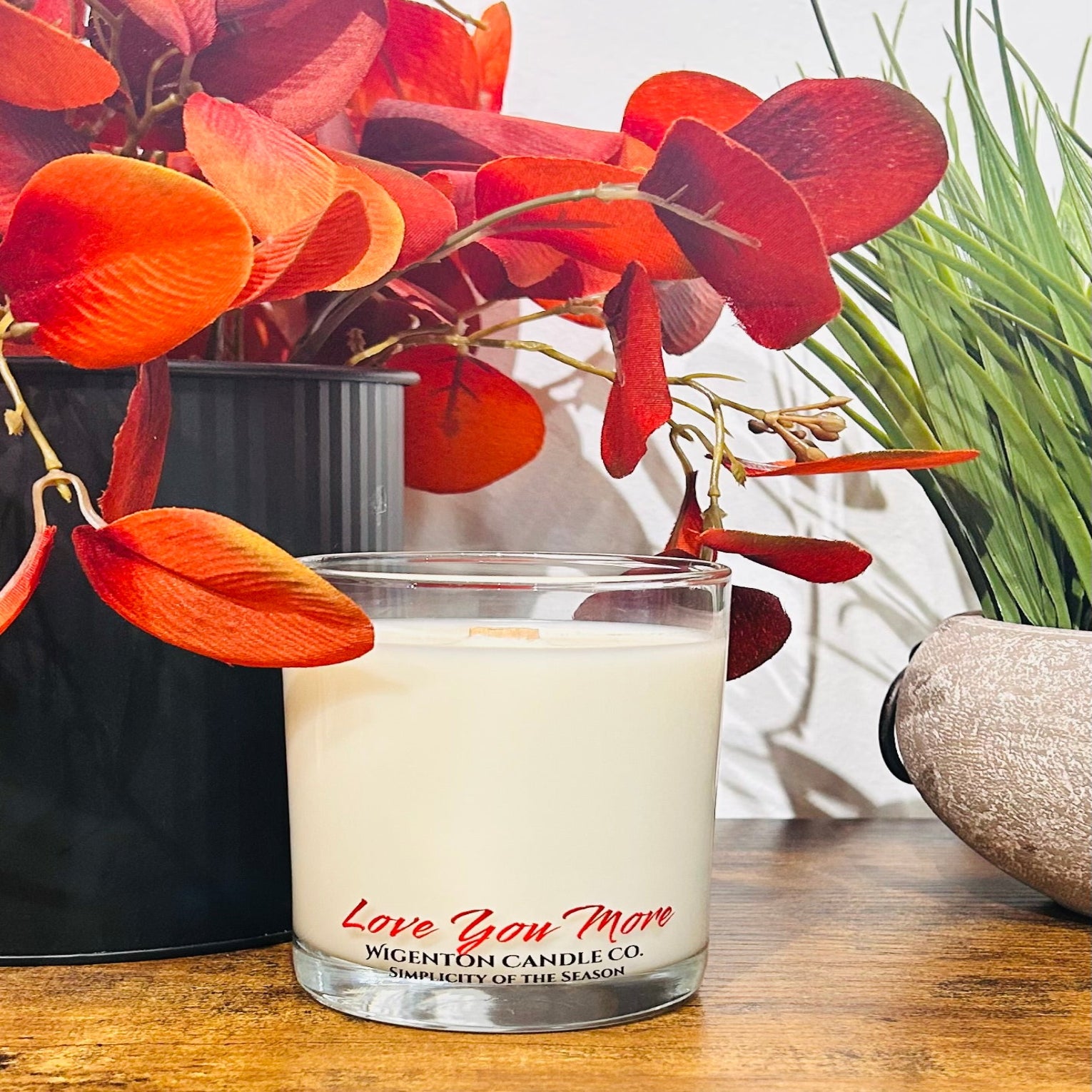 Candle with 'Love You More' text in a glass container on a wooden surface with flowers in the background